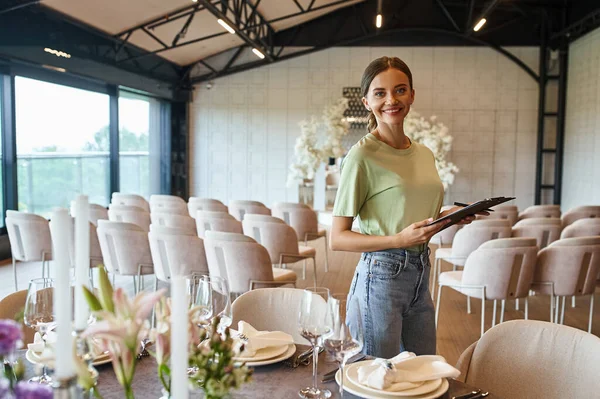 Happy event manager with clipboard smiling at camera near festive table in decorated event hall — Stock Photo