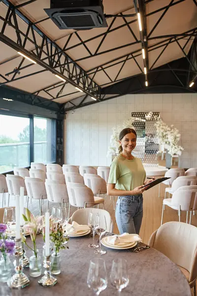Eventmanager mit Klemmbrett lächelt in die Kamera am festlichen Tisch in der Veranstaltungshalle mit floralem Dekor — Stockfoto