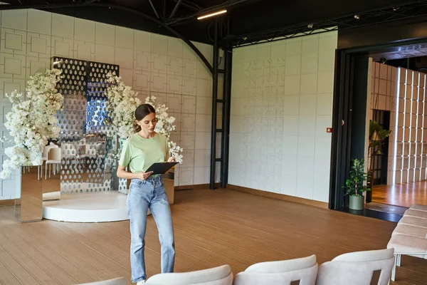 Junger Eventmanager schreibt auf Klemmbrett im modernen Bankettsaal mit Sesseln und floralem Dekor — Stockfoto