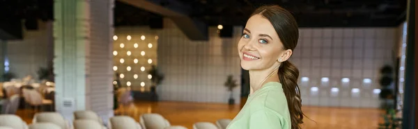 Cheerful creative female decorator looking at camera in spacious and modern event hall, banner — Stock Photo