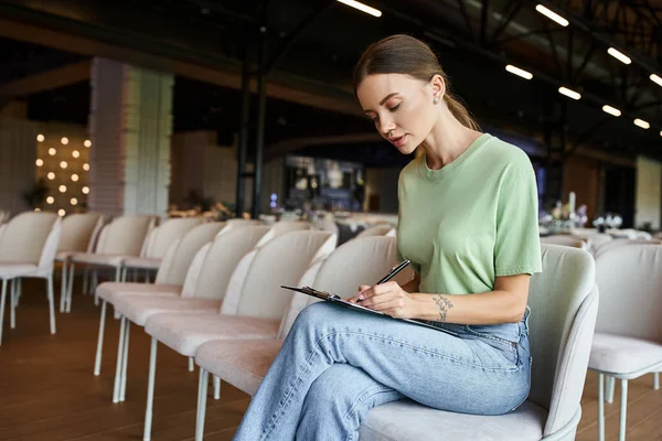 Konzentrierter Eventmanager macht sich Notizen auf Klemmbrett, während er im Sessel im Bankettsaal sitzt — Stockfoto