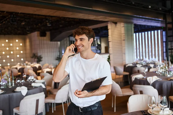 Smiling event organizer with clipboard talking on smartphone near festive tables in banquet hall — Stock Photo