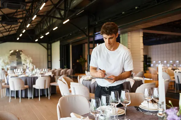 Event decorator making notes on clipboard near tables with festive setting in banquet hall — Stock Photo