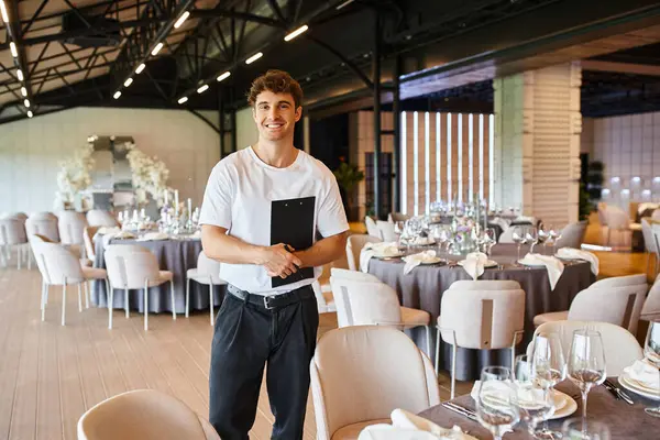 Pleased event manager with clipboard smiling at camera near festive tables in celebration hall — Stock Photo