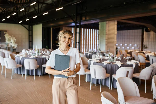 Sorrindo loira gerente de eventos com área de transferência olhando para a câmera no salão de banquetes com mesas decoradas — Fotografia de Stock