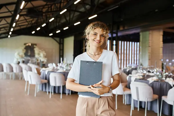 Positive decorator with clipboard looking at camera near tables with festive setting in banquet hall — Stock Photo