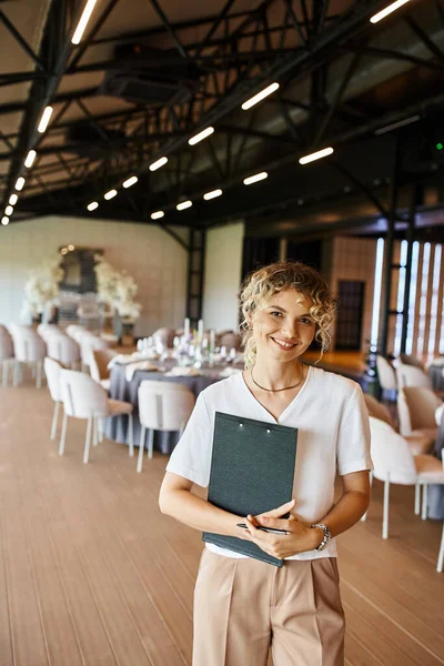 Cheerful event manager with clipboard looking at camera near decorated tables in banquet hall — Stock Photo