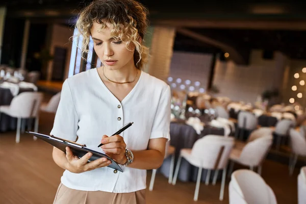 Concentrated event organizer writing checklist on clipboard near festive tables in banquet hall — Stock Photo