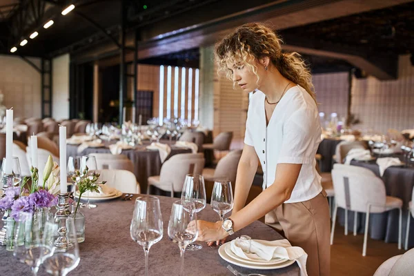 Frau mit welligem Haar stellt Gläser auf festlich gedeckten Tisch, Bankettkoordinator bei der Arbeit — Stockfoto