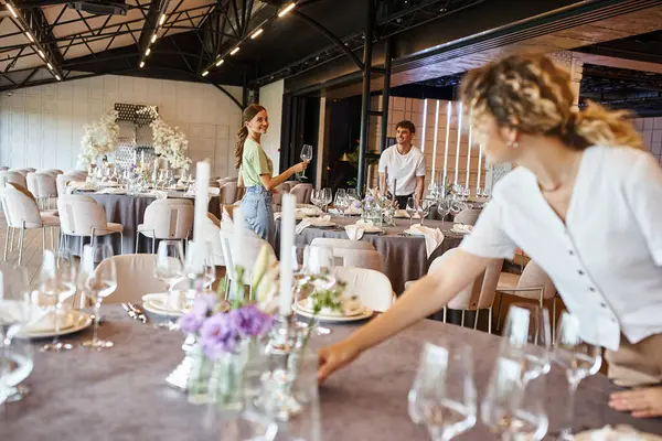 Verschwommene Frau arrangiert festliche Tischdekoration neben lächelndem Team, das in modernem Veranstaltungssaal arbeitet — Stockfoto