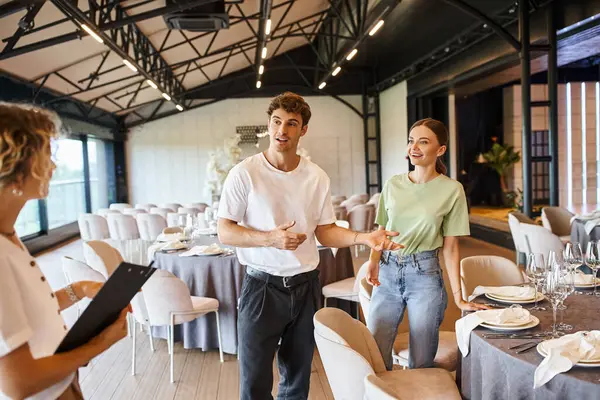 Decorator pointing at table near smiling colleague and team lead with clipboard in event hall — Stock Photo