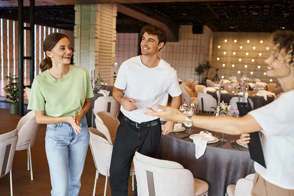 Joyful event manager with clipboard pointing with hand near happy team in modern banquet hall — Stock Photo