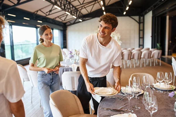 Cheerful man holding plates and arranging festive table setting near creative team in event hall — Stock Photo