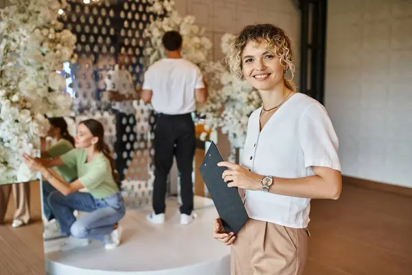 Happy chief decorator looking at camera while team working with floral decor in event hall — Stock Photo