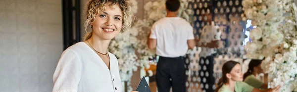 Joyful team lead looking at camera while team of florists working with decor in event hall, banner — Stock Photo
