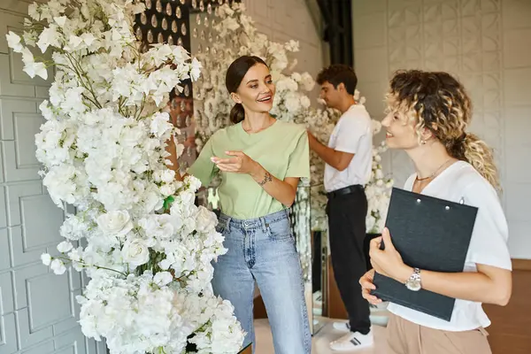 Молодой улыбающийся флорист показывает белый цветочный декор менеджеру мероприятия с буфетом в зале мероприятий — стоковое фото