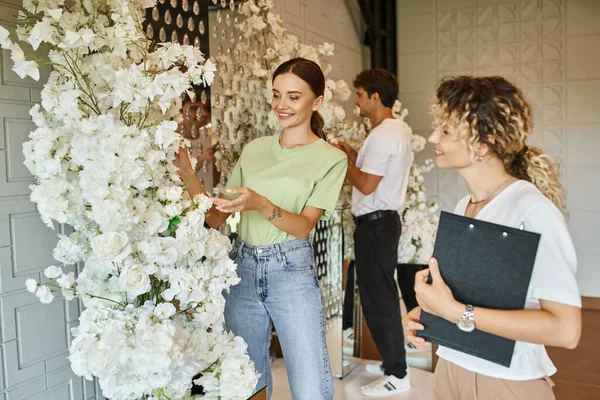 Florista feliz apuntando a blanco floral cerca del equipo de plomo con portapapeles en la sala de eventos, trabajo creativo - foto de stock