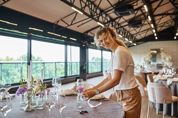 Fiorista felice sorridente alla macchina fotografica e decorare tavolo festivo con fiori per banchetti in sala eventi — Foto stock