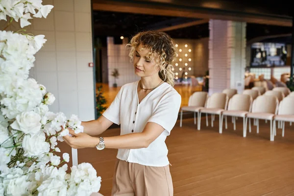 Fiorista bionda con capelli ondulati che organizza composizione floreale bianca in sala eventi, lavoro creativo — Foto stock