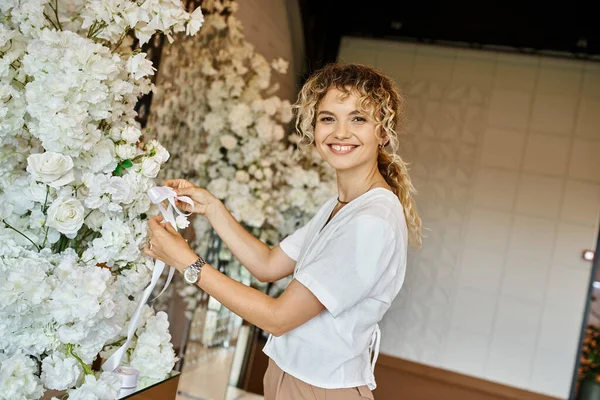Florista alegre decorando salão de eventos com flores brancas e olhando para a câmera, trabalho criativo — Fotografia de Stock