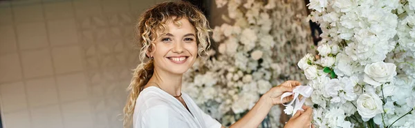 Cheerful florist looking at camera while working with white floral decor in event hall, banner — Stock Photo