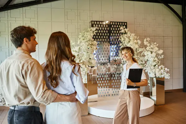 Щасливий менеджер подій з кишені, що показує місце проведення весілля з білою квітковою композицією для пари — стокове фото