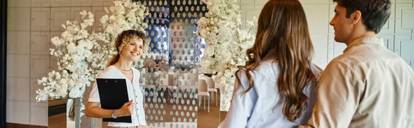 Organizador de eventos com área de transferência sorrindo perto do casal no salão do banquete com decoração floral branca, banner — Fotografia de Stock