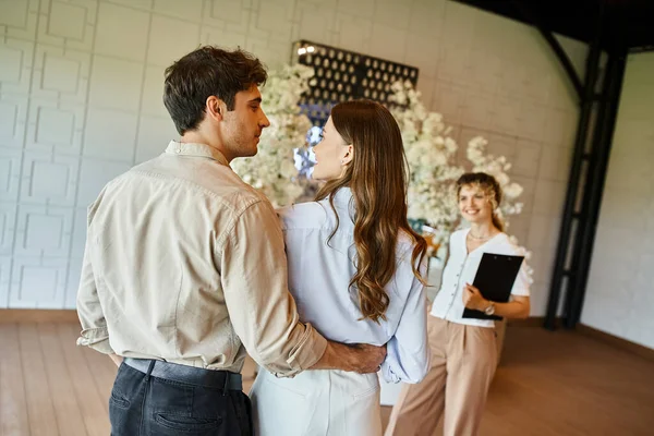 Coppia innamorata guardarsi vicino coordinatore evento nella sala banchetti con decorazione floreale — Foto stock