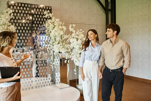 Dekorateur zeigt dem lächelnden Paar weiße Blumenkomposition in der Veranstaltungshalle, Hochzeitsvorbereitung — Stockfoto