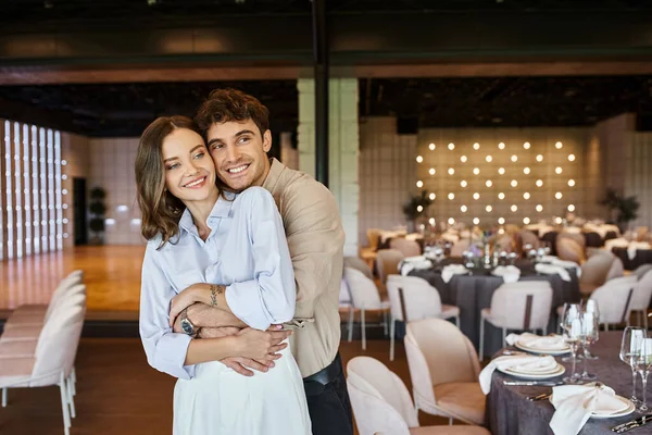 Homem feliz abraçando namorada no salão do banquete com mesas festivas decoradas, configuração do casamento — Fotografia de Stock