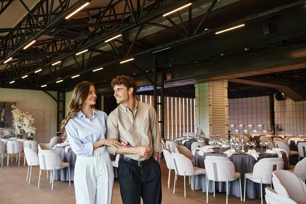 Casal romântico feliz sorrindo um para o outro no salão de banquetes moderno com mesas festivas decoradas — Fotografia de Stock