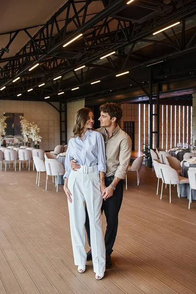 Freudiger Mann umarmt Freundin im geräumigen Bankettsaal mit festlichen Tischen und Hochzeitsdekor — Stockfoto