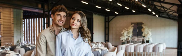 Couple joyeux regardant la caméra dans la salle de banquet moderne et spacieuse avec décor de mariage, bannière — Photo de stock