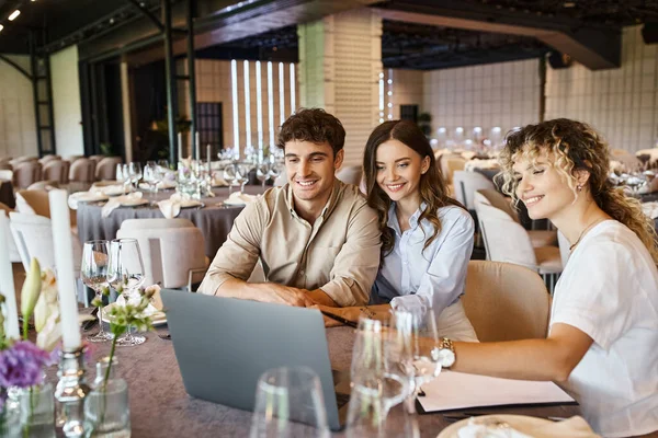 Smiling event manager showing presentation on laptop to happy couple in event hall, wedding setup — Stock Photo
