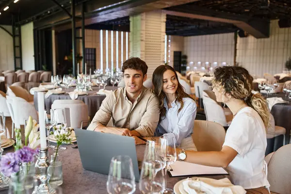 Happy couple in love sitting near banquet coordinator and laptop at festive table in event hall — Stock Photo