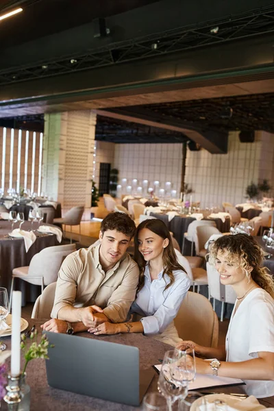 Lächelnder Mann zeigt auf Laptop neben Freundin und Eventmanager am Festtisch in Hochzeitslocation — Stockfoto