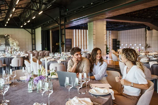 Happy couple and professional event manager talking near laptop at festive table in wedding venue — Stock Photo