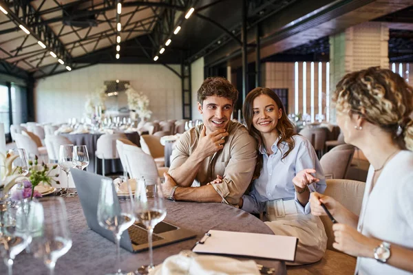 Happy couple and event manager discussing wedding preparation at festive table in banquet hall — Stock Photo