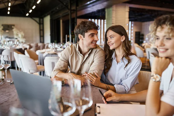 Happy couple talking near blurred event manager near laptop on festive table in banquet hall — Stock Photo