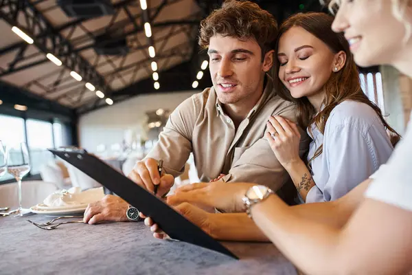 Happy man signing contract near young girlfriend and banquet organizer in decorated wedding venue — Stock Photo