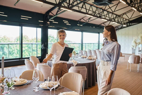 Organizador de eventos com área de transferência apontando para a mesa com ambiente festivo perto da mulher no salão do banquete — Fotografia de Stock