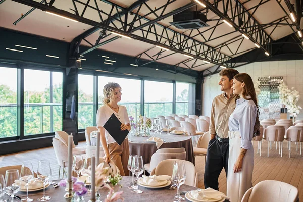 Coppia sorridente innamorata parlando con decoratore di eventi vicino a tavoli festivi in un moderno luogo di nozze — Foto stock
