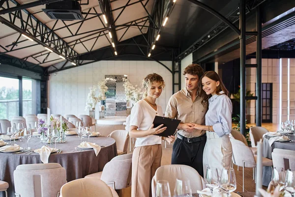 Casal feliz olhando para o contrato na área de transferência perto do coordenador do banquete no salão de eventos decorado — Fotografia de Stock