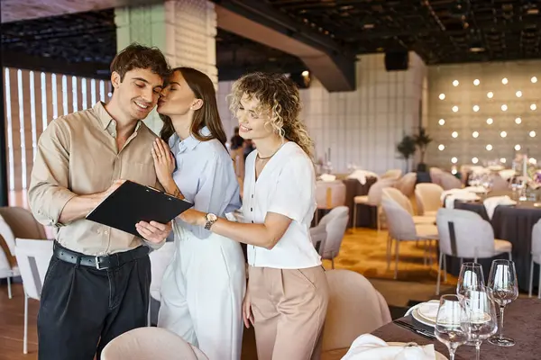 Joyful young woman kissing boyfriend signing contract with event decorator in modern banquet hall — Stock Photo