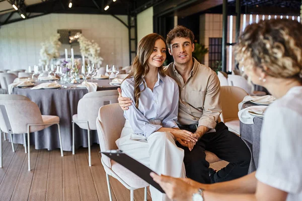 Organizador do banquete com prancheta conversando com casal apaixonado no salão de eventos com decoração de casamento — Fotografia de Stock