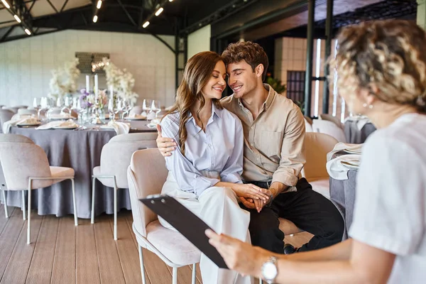 Feliz casal apaixonado sentado com olhos fechados perto do gerente do evento com área de transferência no local do casamento — Fotografia de Stock