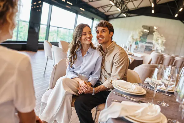 Couple joyeux en amour assis à la table de fête à côté du coordonnateur de l'événement dans le lieu de mariage — Photo de stock
