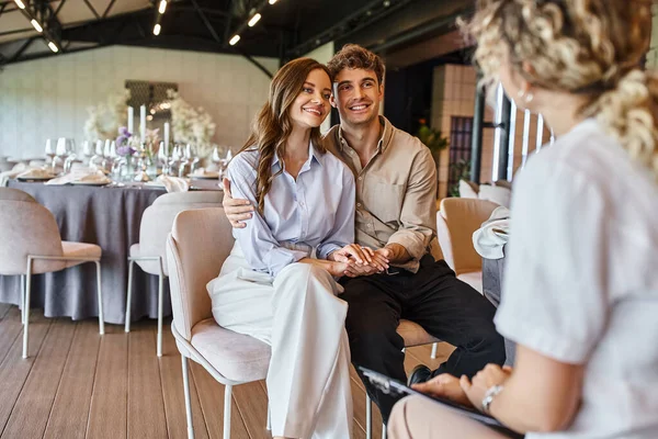 Счастливая пара смотрит на размытого менеджера событий, сидя за праздничным столом в свадебном зале — стоковое фото