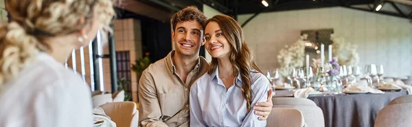Sorrindo casal olhando para gerente de evento turvo e sentado na mesa festiva no salão de casamento, banner — Fotografia de Stock