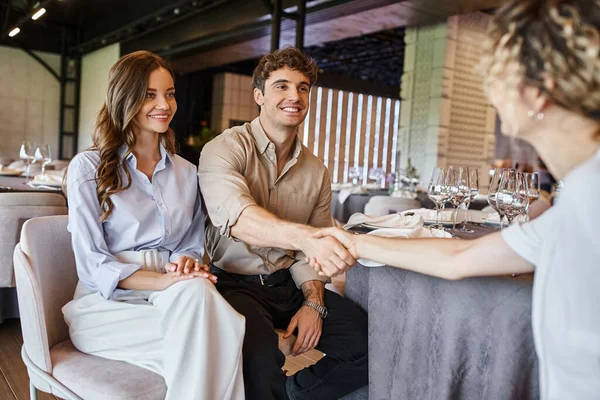 Hombre feliz estrechando la mano con gerente de eventos cerca de novia sonriente en la mesa festiva en la sala de eventos - foto de stock